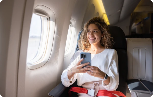 eSIMfirst Roaming - Frau sitzt im Flugzeug am Fenster, lächelt und hält ein Smartphone in der Hand.