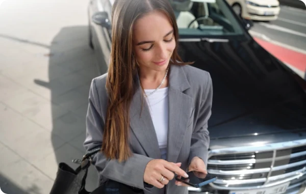 SIXT+ Auto Abo - Geschäftsfrau steht vor einem geparkten Auto und schaut auf ihr Smartphone.