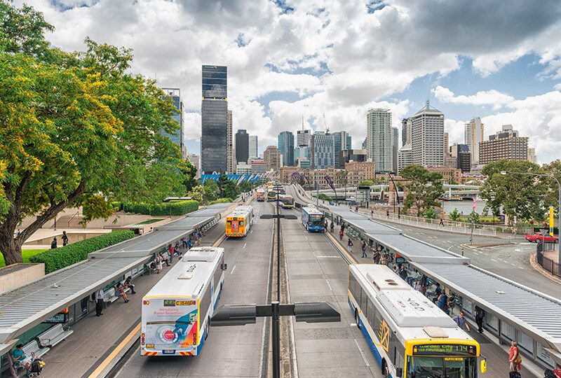 Brisbane busway with city skyline in the background and commuters at platforms.