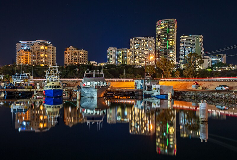 Darwin city skyline at night with boats reflected in the calm harbour.