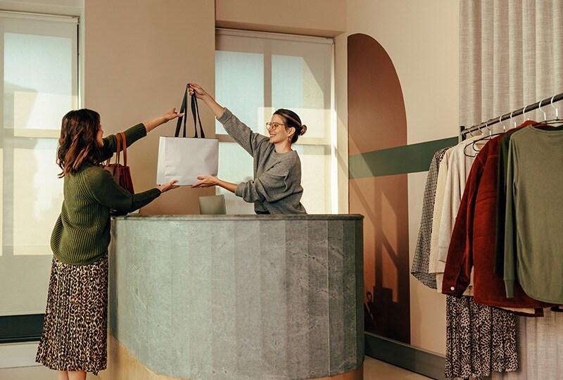 Shop assistant handing a shopping bag to a customer inside a boutique clothing store.