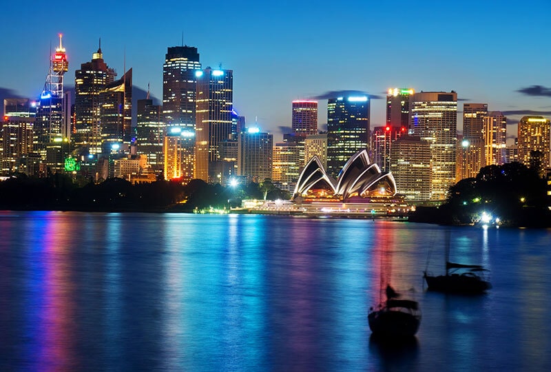 Sydney Opera House and city skyline illuminated at night, reflecting across the harbour.