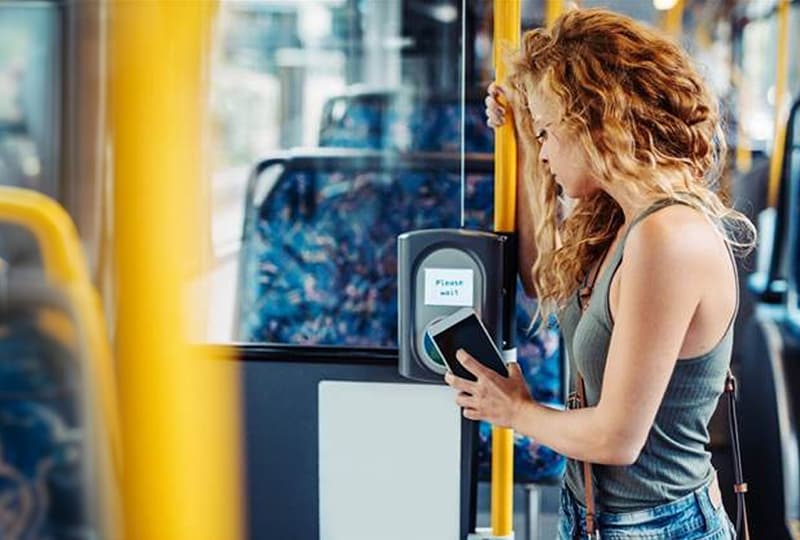 Woman holding a smartphone to a contactless fare reader on a city bus, standing by yellow poles and patterned seats; the reader’s screen says “Please wait.”