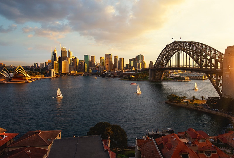 Sydney Harbour at sunset with the Opera House, Harbour Bridge, and sailing boats.