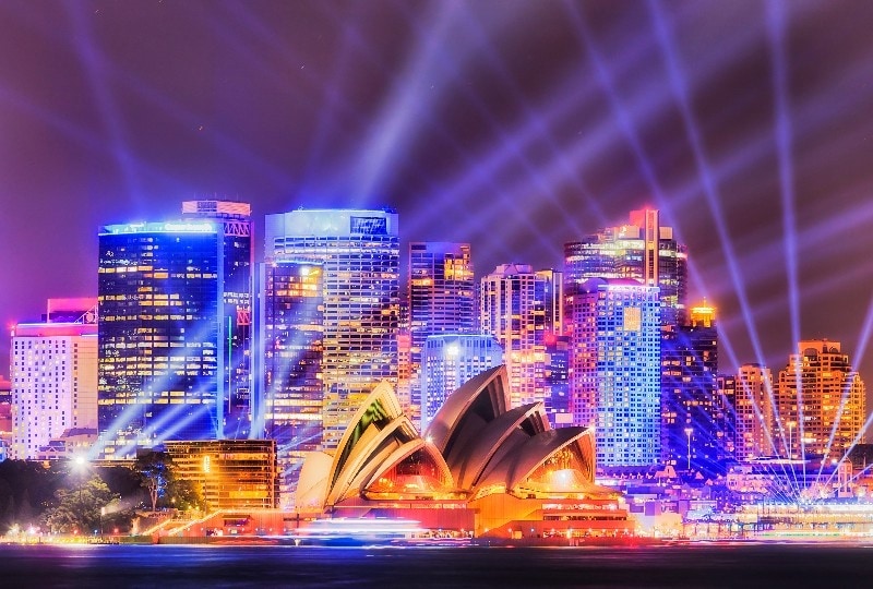 Sydney Opera House and city skyline illuminated with colourful light beams at night.
