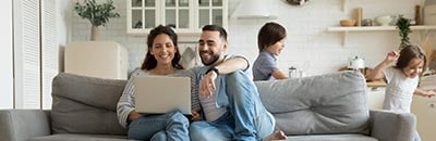 couple using laptop on living room couch as kids run and play