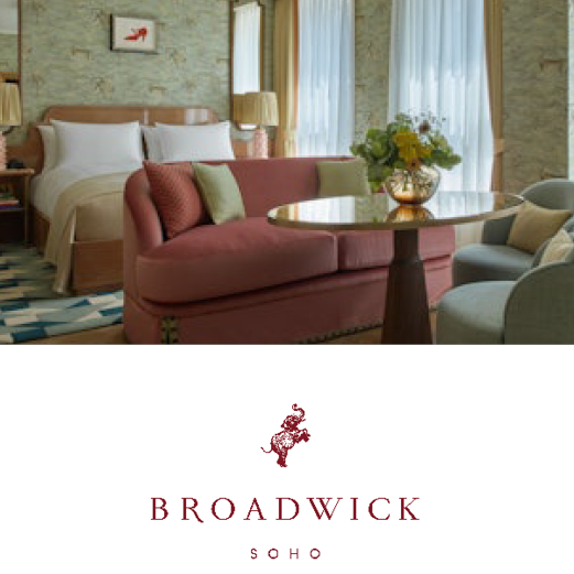 Broadwick, Soho. A luxurious Broadwick hotel room. A red velvet sofa sits at the foot of the double bed. A vase of fresh flowers sits on a table and sun shines through the window.