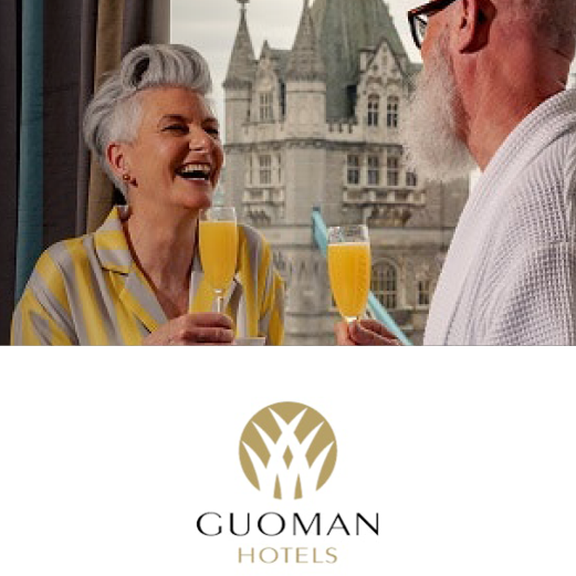 Guomon Hotels. A happy and laughing couple stand facing each other. They are holding a glass of orange juice and champagne. The window behind looks out upon Tower Bridge.