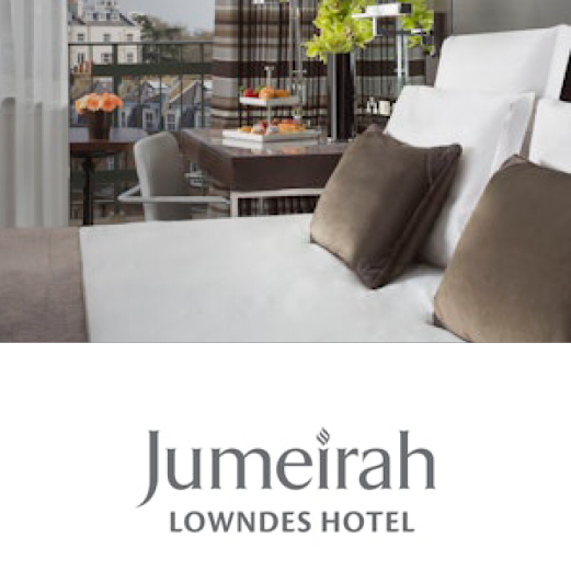 Jumeirah, Lowndes Hotel. A closeup view of a modern hotel room. Themed in whites and greys. A table sits in the background by the window with a vase of flowers and bowl of fruit upon it.