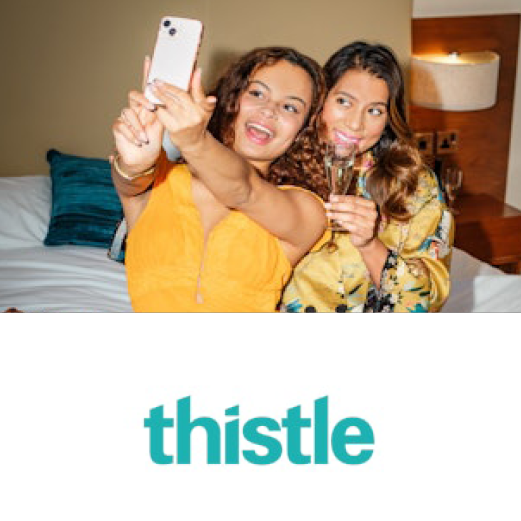 Thistle. Two young women pose for a selfie in their hotel room. One holds her phone out in front to take the photo and the other smile holding a glass of champagne.