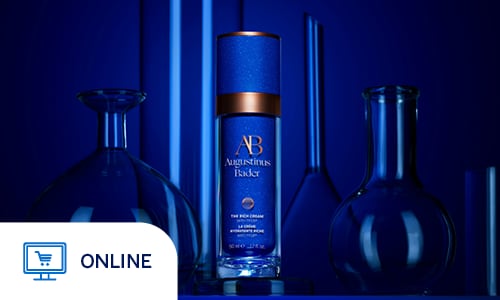 A sleek blue bottle of Augustinus Bader "The Rich Cream" stands at the center of a scientific and sophisticated display. The product features a deep blue body with copper-toned accents on the cap and logo, positioned on a clear circular pedestal. Surrounding the bottle are various glass laboratory vessels, including round-bottom flasks and cylinders, all bathed in a dramatic monochromatic blue light. The composition uses deep shadows and glowing highlights to emphasize a laboratory-inspired, high-end aesthetic, suggesting a blend of luxury and clinical science. Online Only.