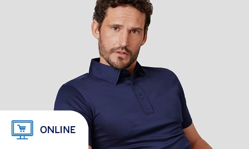 A man with curly dark hair and a light beard is pictured sitting in a green chair against a plain grey background. He is wearing a dark navy blue polo shirt with a classic collar and a two-button placket, paired with khaki-colored trousers. He is posed leaning slightly forward with a neutral expression, creating a clean and professional studio portrait that emphasizes a smart-casual menswear aesthetic. Online only.