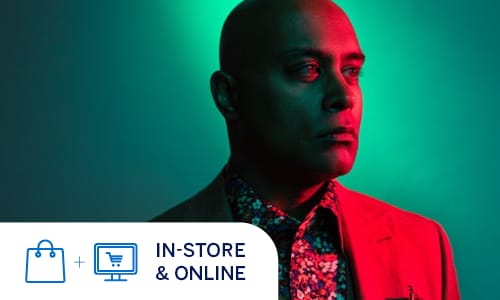 A man with a shaved head is captured in a striking, cinematic portrait lit by vibrant red and teal light. He is dressed in a tailored three-piece suit consisting of a light brown or grey blazer and matching vest, paired with a colorful, floral-patterned button-down shirt. A patterned pocket square peeks out from his breast pocket, adding another layer of detail to his ensemble. He is looking off-camera with a contemplative expression, and the dual-tone lighting creates a high-contrast, artistic effect that highlights the contours of his face and the textures of the fabric. In-store and online.