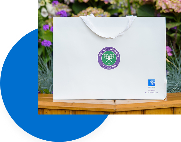 A white bag with The Championships, Wimbledon logo in the center and the American Express logo in the bottom right sitting on a wood rail in front of flowers.