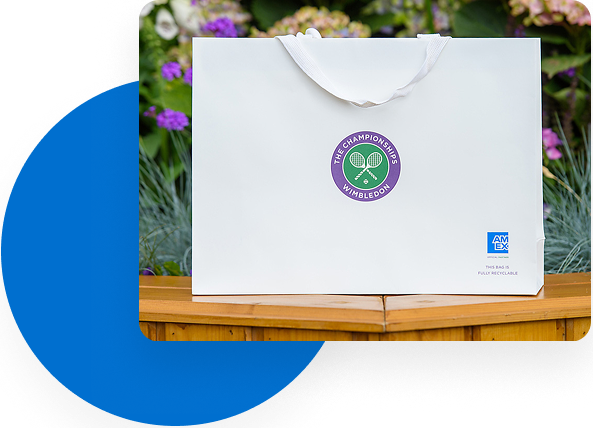A white bag with The Championships, Wimbledon logo in the center and the American Express logo in the bottom right sitting on a wood rail in front of flowers.