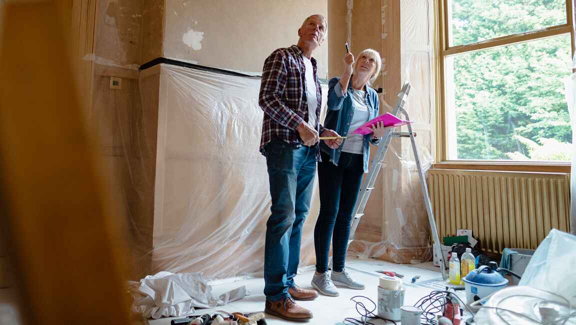 Couple are surrounded by decorating equipment in their home, pointing to the ceiling with a pencil