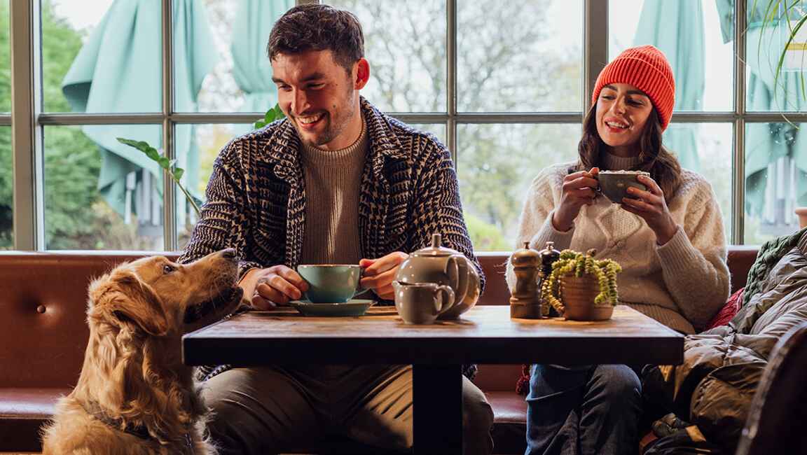 Two friends are smiling at a dog while sat drinking tea in a cafe
