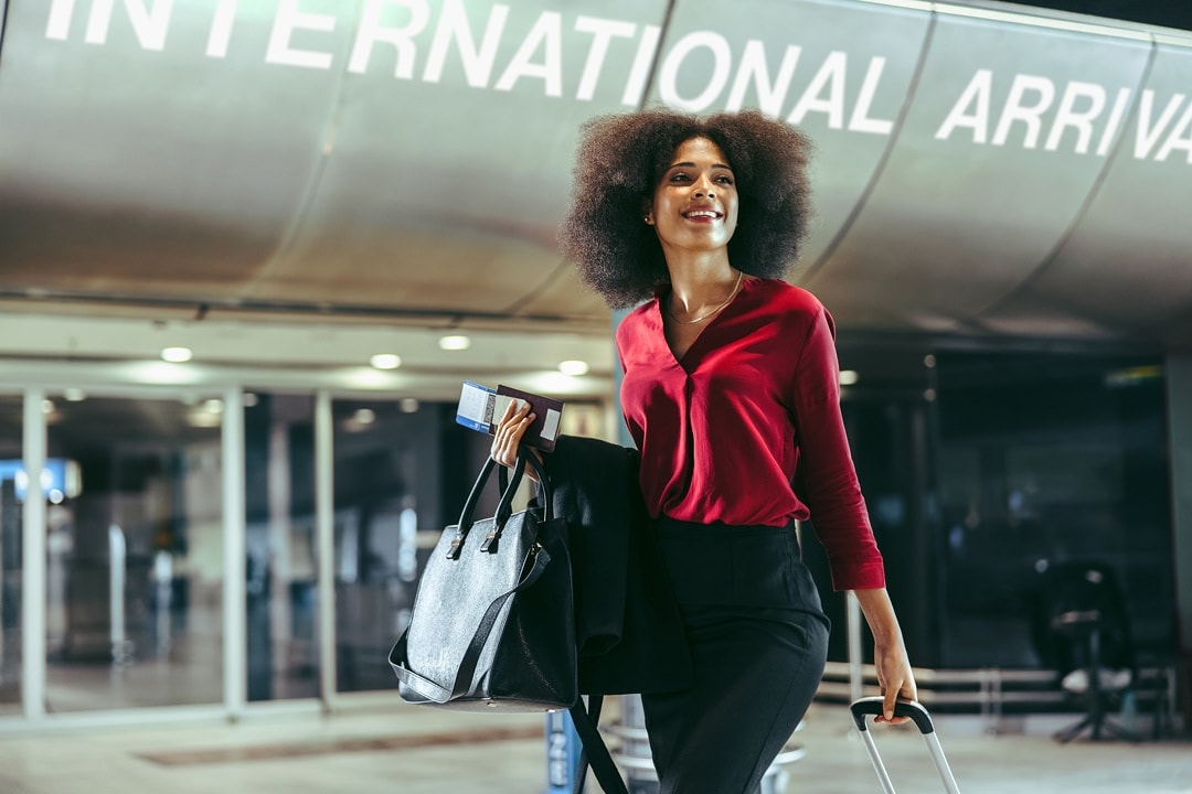 Woman walking through an airport arrivals area with a suitcase and passport