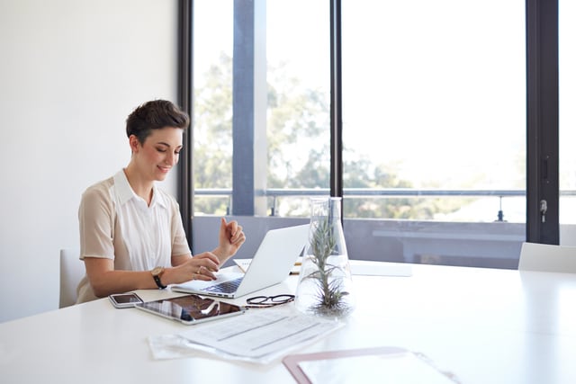 Businesswoman typing on laptop in meeting room, tablet, phone & glasses on table.