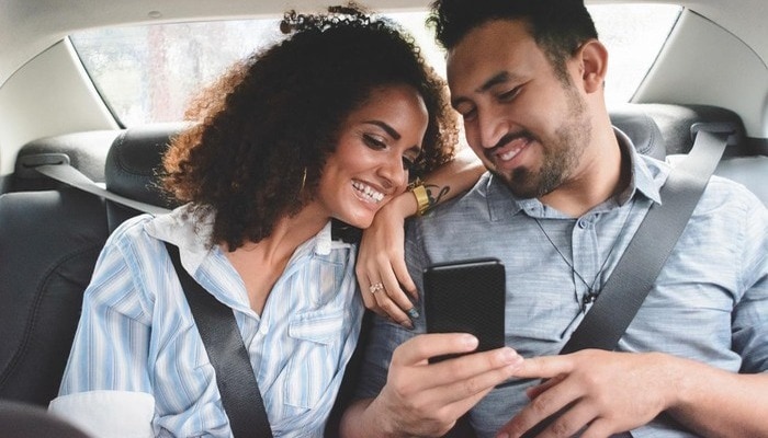 A couple in the back of a car, smiling whilst looking at a mobile phone.