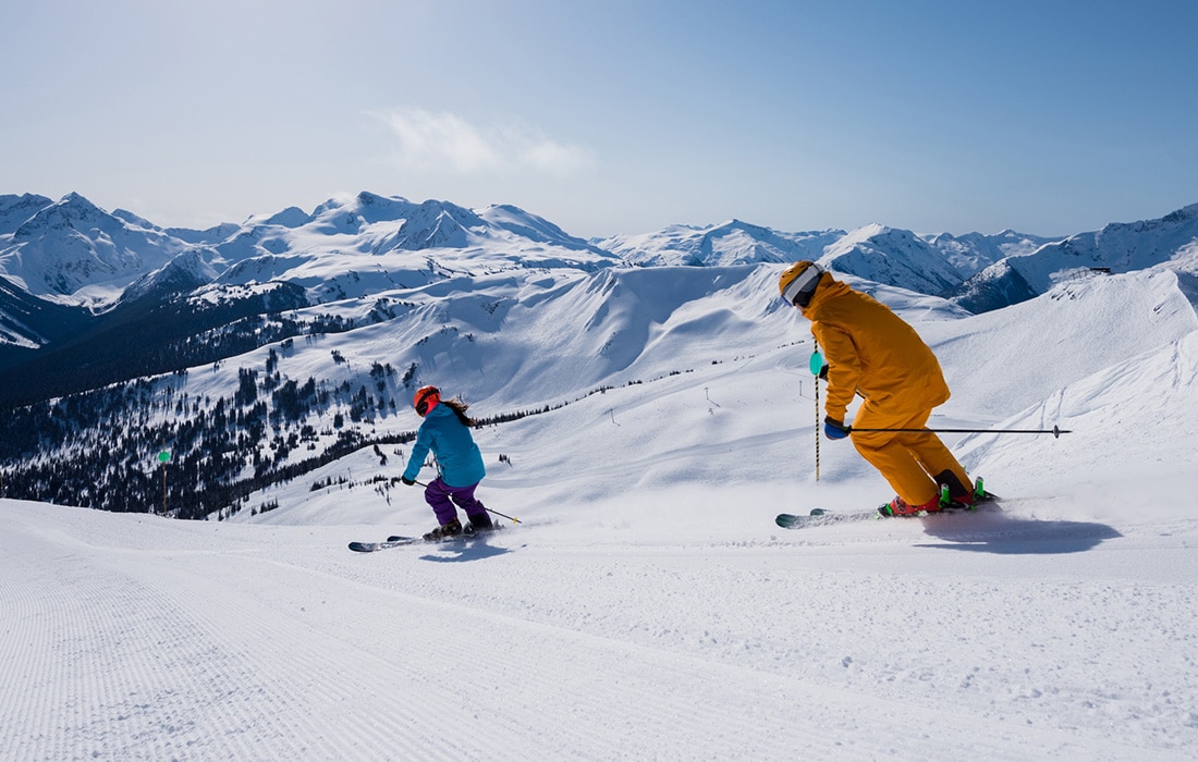 Two people skiing down a snowy slope in the mountains.