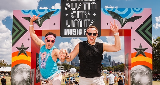 Two people jumping in front of a colorful ACL Festival sign