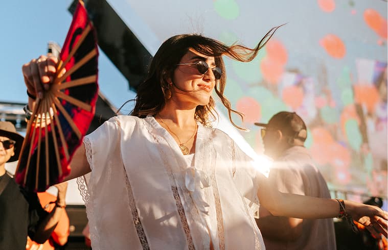 Festival attendee holding a fan dances in a crowd, with colorful lights in the background.