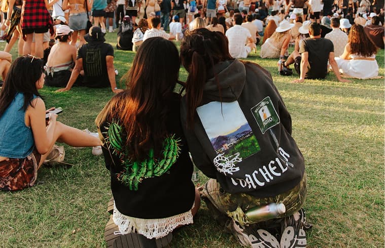 Two festival attendees sit together on the grass, wearing Coachella merchandise.