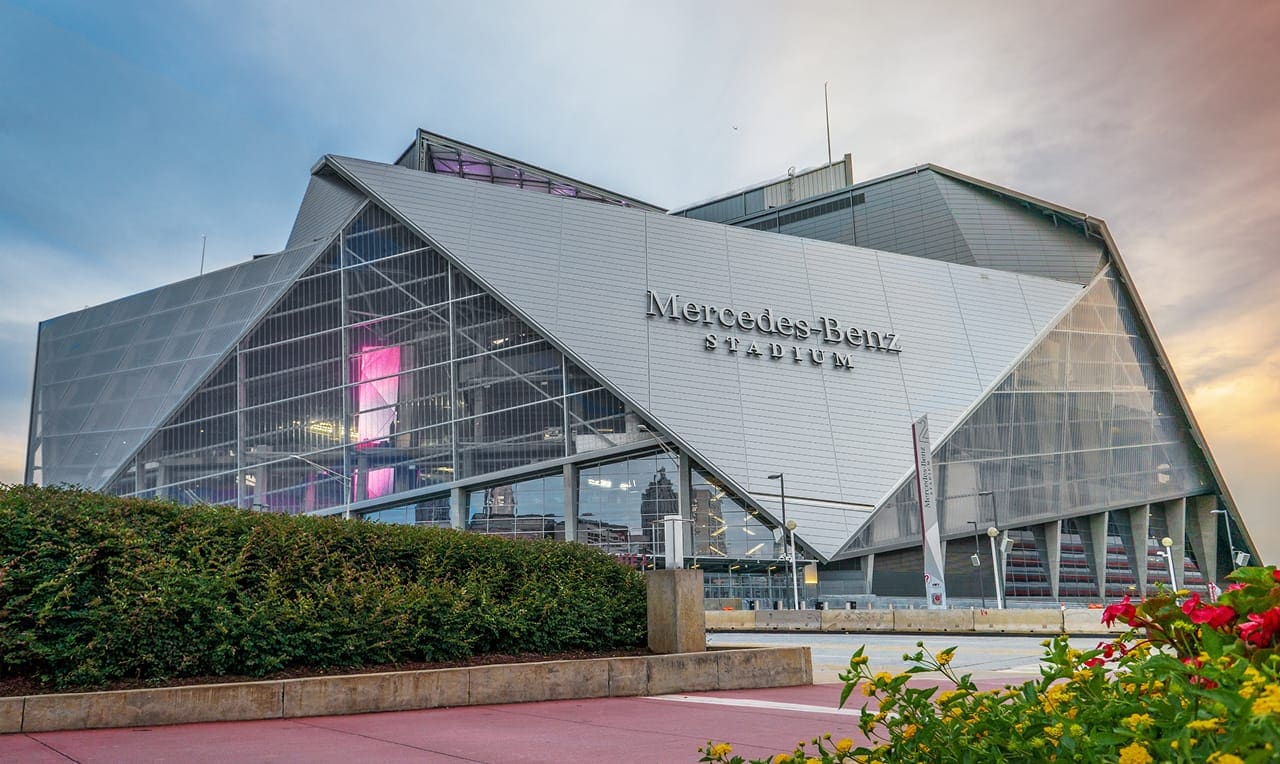 A photograph of the Mercedes Benz Stadium, with a cloudy sky and the sunset in the background.