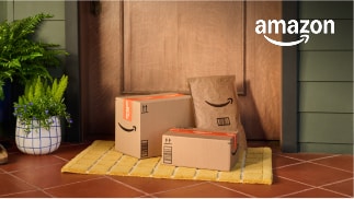 Three Amazon packages set in front of a wooden door, on top of a yellow welcome mat, and to the right of a potted plant.