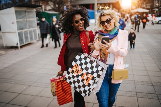 Two women with sunglasses on carrying shopping bags, walking and laughing going down the street