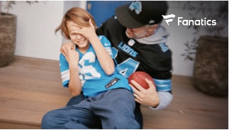 Photo of a son on a dad's lap laughing in Lions gear. The dad is holding a football laughing. 