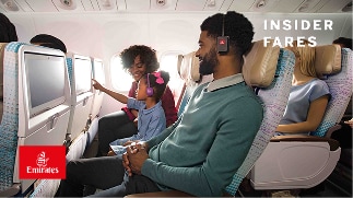A dad in a green sweater sitting next to his daughter on the plane. The daughter is touching the screen on the seat in front of her as her mom watches.