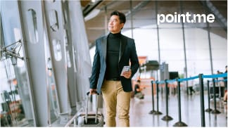 A man in tan pants and dark suit jacket walking in the airport pulling his suitcase and holding his phone / boarding pass.