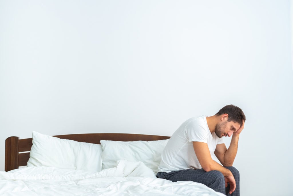 Man sitting at edge of bed with head in hand looking disturbed.