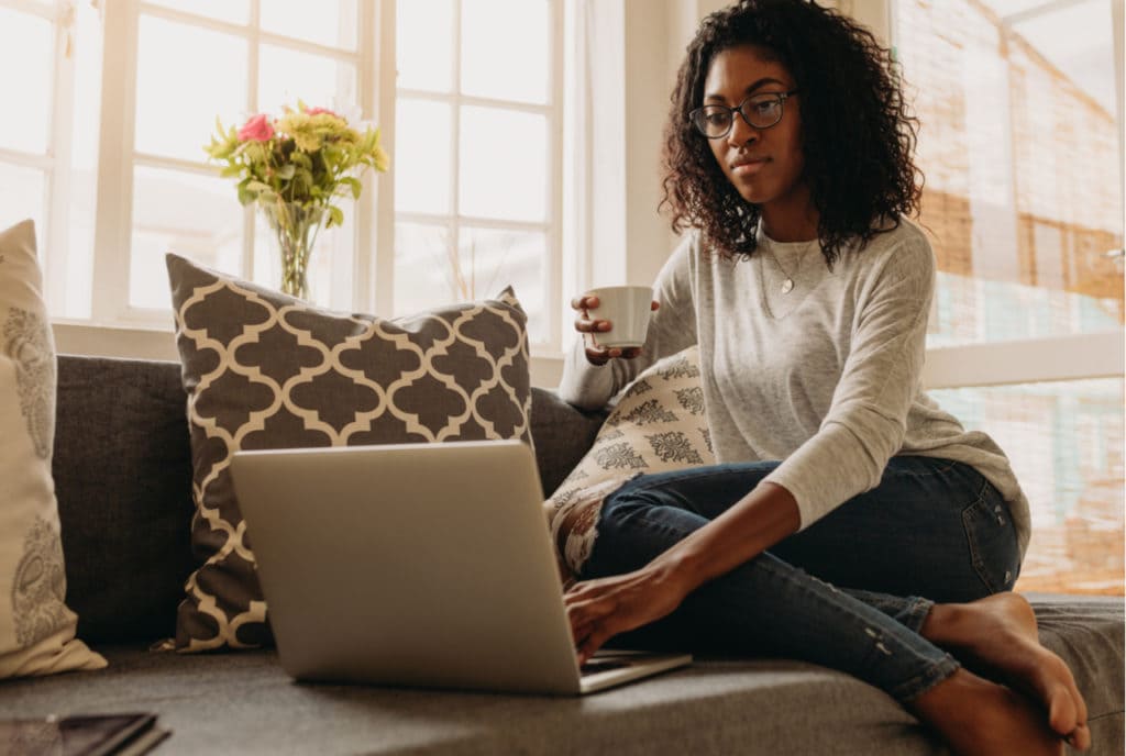 Woman sitting on couch with cup in hand looking at computer.