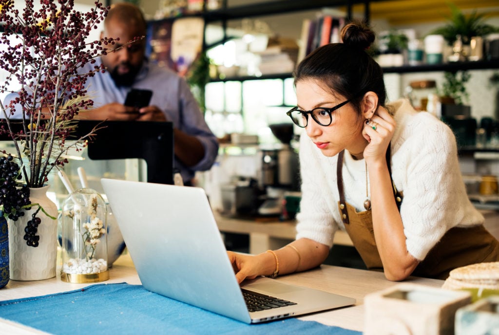Woman with glasses looking at computer in coffee shop while man is looking at phone behind her.