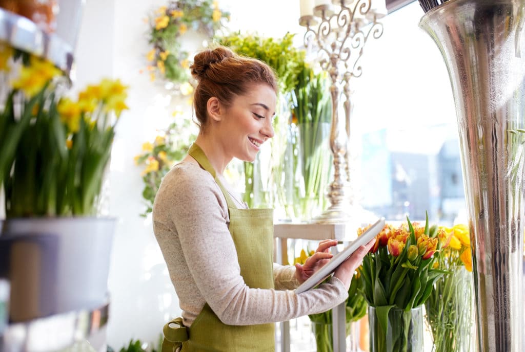 Woman standing in flower shop looking at ipad while smiling.