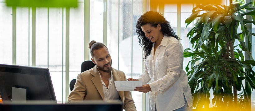 A businesswoman meets with a businessman in his office showing him some paperwork while he is seated at his desk.