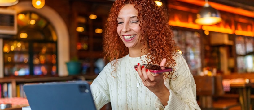 A woman sitting in a cafe holds her phone while checking her tablet