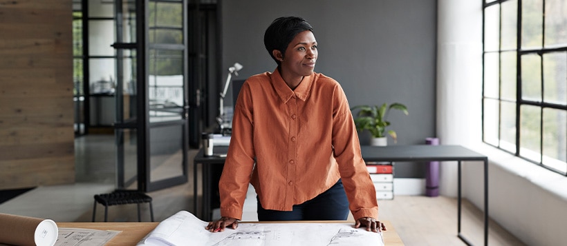 While in her office a woman stands over architecture maps while staring out of the window