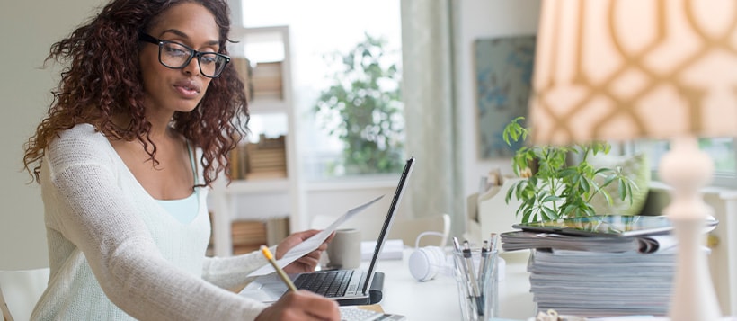 A woman in glasses reviewing paperwork and taking notes at her desk in front of her laptop.