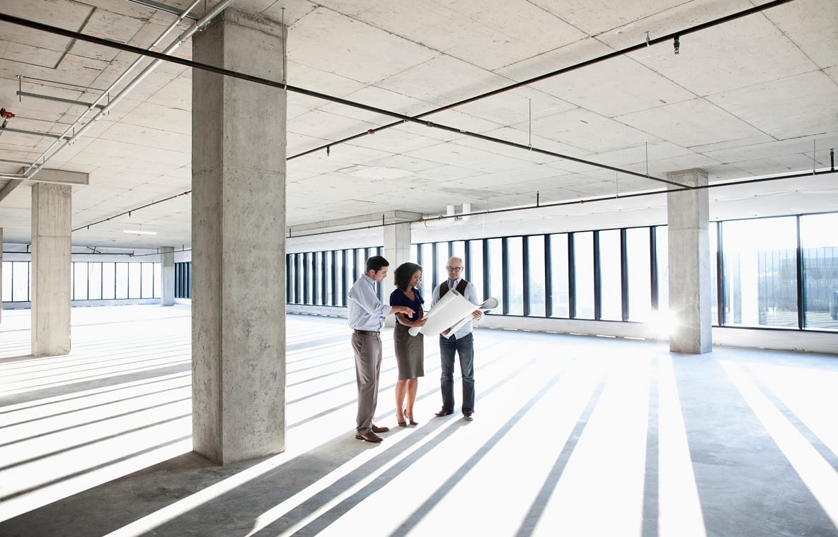 Three professionals reviewing blueprints in an empty, sunlit office space