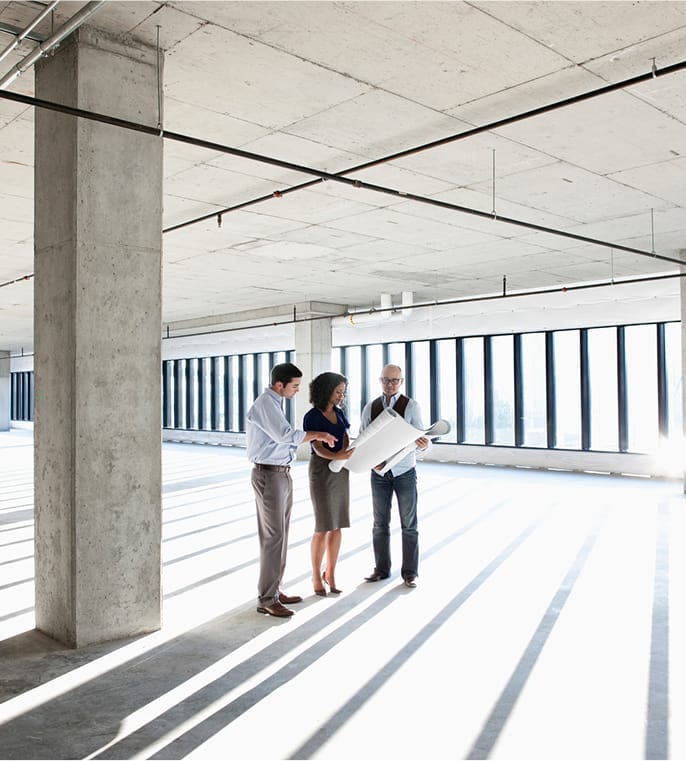 Three professionals reviewing blueprints in an empty, sunlit office space