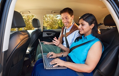 Woman typing on a laptop from backseat of a car next to a colleague.