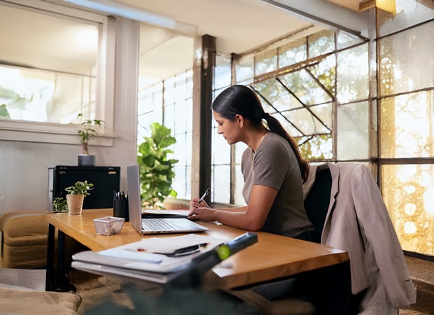 A businessperson sits at their desk writing with a pen and paper, with a laptop on the desk and plants around the space.