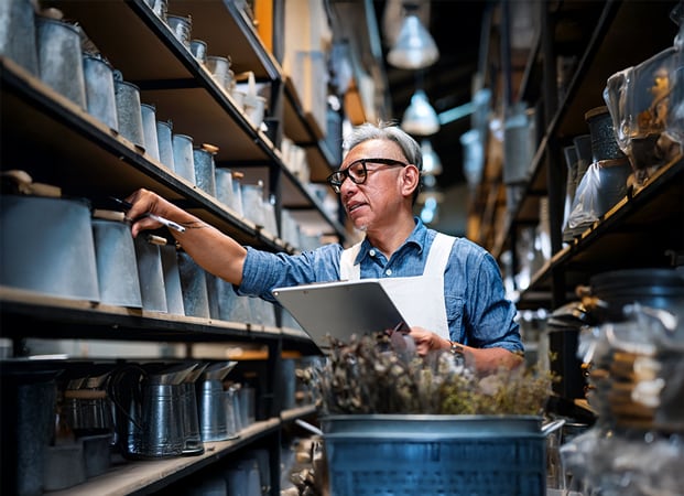 A businessperson sorts through metal buckets on a shelf in an industrial workspace while holding a tablet.