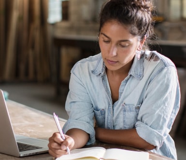 A woman in business-casual clothes writes in a notebook with her laptop open for reference.