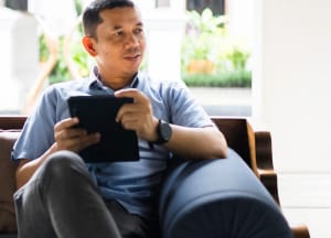 A man in business-casual clothes holds a discussion on a couch while holding his tablet.