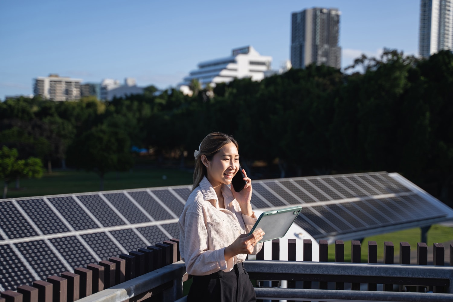 A businesswoman out on the field reports on her phone, holding her tablet up for reference.